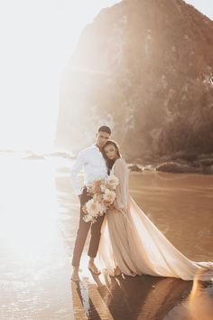 a bride and groom standing on the beach with their arms around each other, posing for a photo