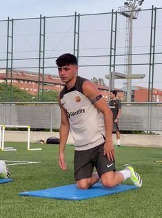 a young man stretching on a blue mat in the middle of a soccer field with other players behind him