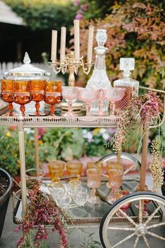 a table with glasses, candles and flowers on it in the middle of a garden