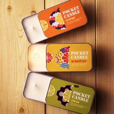 three candles in tins sitting next to each other on a wooden surface, one with an orange and the other with a pocket candle