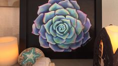 a painting of a blue succulent in a black frame next to candles and seashells