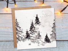 a card with watercolor trees on it sitting next to string lights and christmas lights