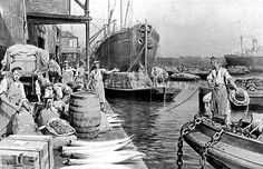 an old black and white photo of people loading cargo onto boats at a dock with ships in the background