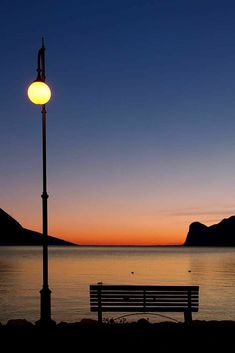 a bench sitting next to a lamp post near the ocean at sunset or dawn with mountains in the background