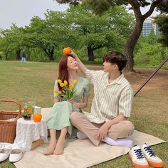 a young man and woman sitting on a blanket in the park with flowers, oranges and fruit