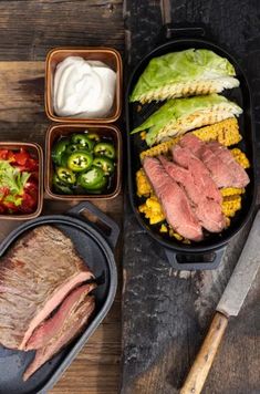 an assortment of meats and vegetables in trays on a wooden table with utensils