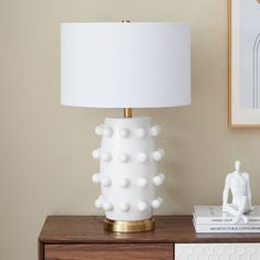 a white lamp sitting on top of a wooden dresser next to a framed painting and a book