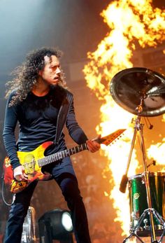 a man with long hair playing guitar in front of a large fire and flames behind him