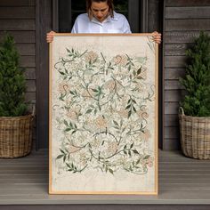 a woman holding up a large framed painting on the front porch with potted plants behind her