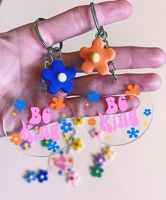 a hand holding two clear round keychains with colorful flowers on them and the words be kind written in large letters