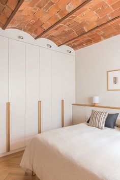 a bed sitting under a wooden ceiling next to white cupboards and a lamp on top of a hard wood floor