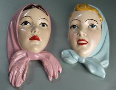 two ceramic heads are sitting next to each other on a gray surface, one has blue eyes and the other has blonde hair