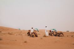 two men on camels in the desert with trucks behind them and people standing around