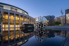 an artistic sculpture sits in front of a building at night with its reflection on the water