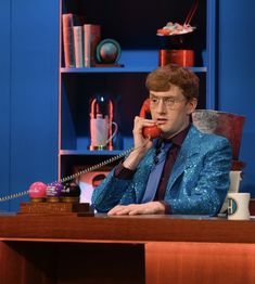 a man sitting at a desk talking on a phone while wearing a suit and tie