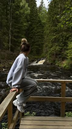 a woman is sitting on a wooden bridge looking at a river in the woods and trees