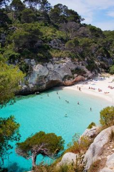 people are on the beach and in the water near some trees, rocks and sand