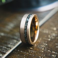 a wedding band with black and gold inlays on it sitting on top of a wooden table