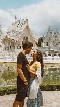 a man and woman standing next to each other in front of a lake with white buildings behind them