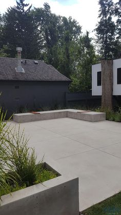 an empty concrete patio surrounded by greenery and trees in front of a gray house