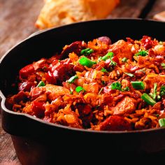 a skillet filled with rice and meat on top of a wooden table next to bread