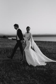 a bride and groom walking in the grass by the water on their wedding day, black and white photo