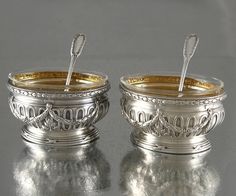 two silver cups with spoons in them on a reflective surface, one is empty