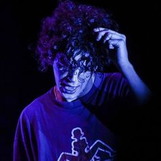 a man with curly hair wearing a t - shirt and holding his hand to his head