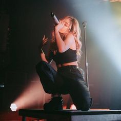 a woman sitting on top of a stage holding a microphone in her hand and wearing black pants