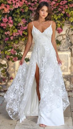 a woman wearing a white dress with flowers on it and a slited skirt is standing in front of a stone wall