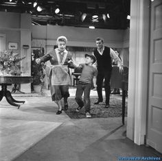 an old black and white photo of three people walking in front of a tv screen