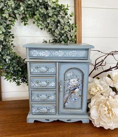 a blue dresser with flowers and a mirror in the background on top of a wooden table