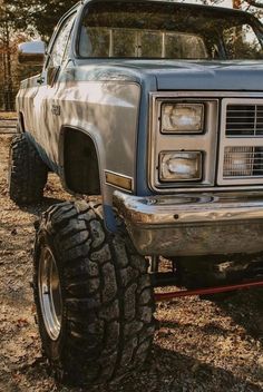 the front end of a silver truck parked on top of a dirt field with trees in the background