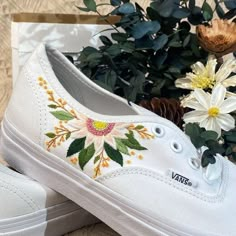 a pair of white shoes with embroidered flowers on them next to some plants and pine cones