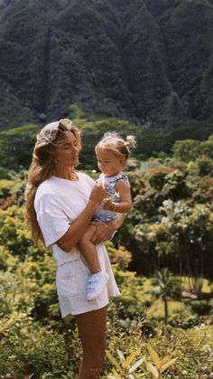 a woman holding a baby in her arms while standing on top of a lush green hillside