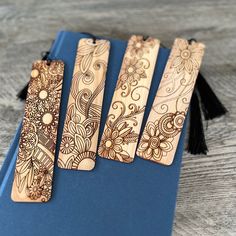 four wooden bookmarks with designs on them sitting on a blue binder next to a black tassel