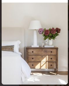 a white bed sitting next to a wooden dresser with flowers on top of it in a bedroom