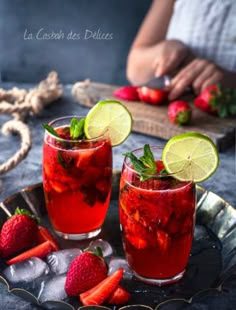 two glasses filled with red liquid and strawberries on a metal tray next to a woman