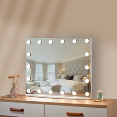 a bedroom with a dresser and mirror that has lights on the top, and a bed in the background