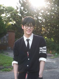 a young man wearing glasses and a black sweater is standing in front of some trees