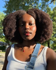 a woman with an afro wearing a white tank top and blue jeans suspenders standing in front of a tree