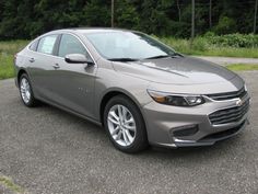 a silver car parked in a parking lot next to some grass and trees on the side of the road