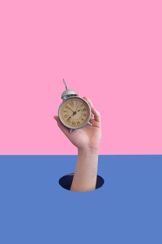 a hand holding an alarm clock in front of a pink and blue background