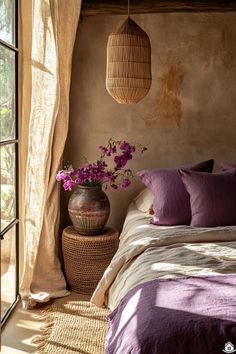 a bed with purple sheets and pillows next to a vase filled with flowers on top of it