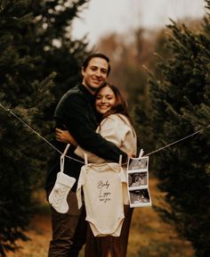 a man and woman standing next to each other in front of christmas trees holding baby onesuits