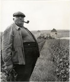 an old man standing in the middle of a field with a pipe sticking out of his mouth