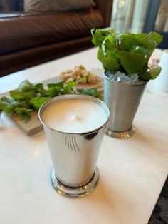 two silver cups filled with plants on top of a white marble table next to a couch