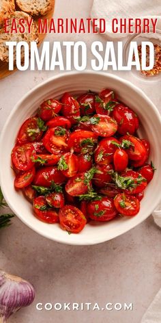 a white bowl filled with tomatoes next to garlic and bread on top of a table