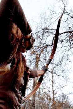 a woman is holding her bow and aiming it at the sky with trees in the background