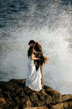 a bride and groom standing on rocks near the ocean with splashing water behind them